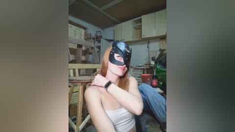 A video of a fair-skinned, red-haired woman wearing a black Batman mask and a white tank top, sitting in a cluttered workshop with wooden furniture and tools in the background.