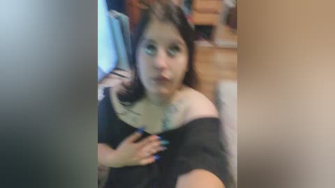 A blurry video of a young woman with fair skin and dark hair, wearing a black off-shoulder top, holding a phone, in a cluttered room with wooden furniture.