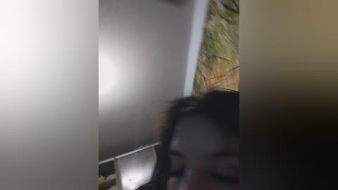 A blurry video shows a young person with dark hair, partially obscured, holding a smartphone near a window with greenery outside.