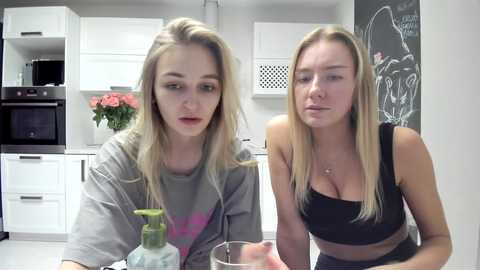 Media: Video of two young women with long blonde hair, one in a grey tee, the other in a black crop top, sitting at a kitchen table, discussing.