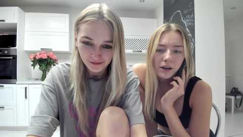 Media: Video of two young women, one blonde in a grey shirt, the other in a black top, sitting closely in a modern kitchen with white cabinets, oven, and a vase of pink flowers.