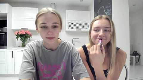 Media: Video of two young women in a modern kitchen. One with blonde hair, wearing a gray graphic T-shirt, sits. The other, also blonde, brushes her teeth, wearing a black top. White cabinets, blackboard, flowers, and sleek appliances in background.