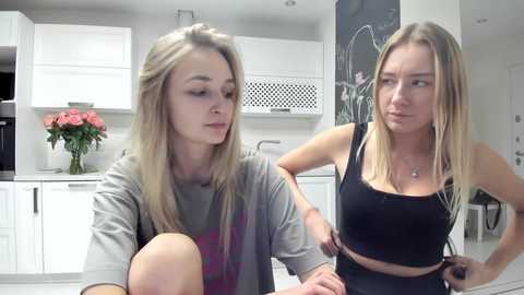 Media: Video of two young women, one with blonde hair, wearing a gray hoodie, the other with blonde hair, wearing a black crop top, standing in a modern kitchen with white cabinets and a blackboard wall.