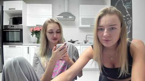 Media: Video of two young Caucasian women with long blonde hair, one wearing a gray top, the other a black tank, drinking coffee in a modern, white kitchen with stainless steel appliances and a microwave.