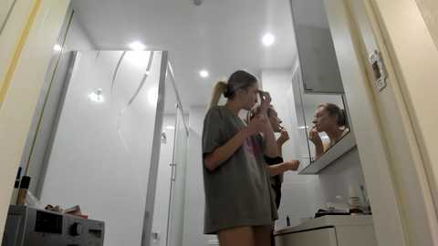 Media: Video of a bathroom with two women; one applying makeup in front of a mirror, another brushing her hair. Modern, white-tiled bathroom with a shower and sink.