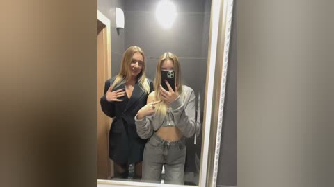 Media: Video of two blonde women, one in a black dress, the other in a gray crop top and pants, taking selfies in a dimly lit bathroom with a large mirror.