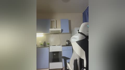 Video of a small, dimly lit kitchen with light blue cabinets, a stove, and a white fridge, featuring a robot vacuum cleaner in the foreground, with a blue curtain on the right.