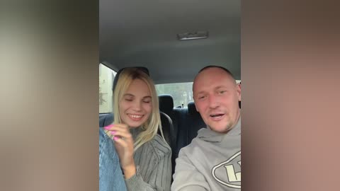 Media: Video of a smiling, blonde Caucasian woman with straight hair, wearing a blue sweater, and a bald Caucasian man in a gray hoodie, sitting in a car.