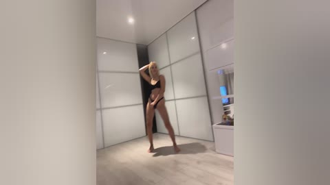 Media: Video of a slender, blonde woman in black lingerie performing a provocative dance in a modern, minimalist bedroom with light wood flooring and frosted glass closet doors.