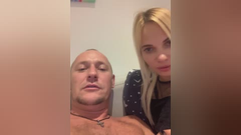 Media: Video of a bald, muscular, shirtless man with a neutral expression next to a blonde woman with a choker, wearing a dark top, in a dimly lit room.