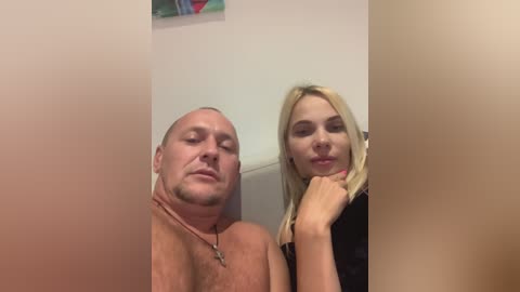 Media: Video of a bald, shirtless, overweight man and a blonde woman with long hair, both smiling, in a bathroom with beige walls and a colorful painting.