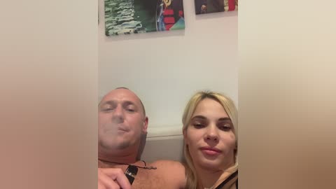 Media: Video of a bald, muscular, light-skinned man and a blonde woman with fair skin, both puffing on cigarettes, seated in a dimly lit, white-walled room adorned with colorful abstract art.