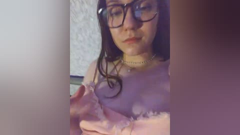 Video of a fair-skinned woman with long brown hair, wearing glasses, a choker, and a lavender top, holding a pink, frosted cake, against a blurred, textured background.