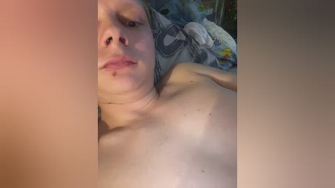 Video of a topless woman with fair skin and wet hair, lying on a bed with a patterned blanket. She has a neutral expression and a small mole on her left shoulder.