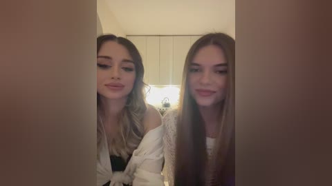 Media: Video of two young Caucasian women with long, straight brown hair, wearing white off-shoulder tops, standing side by side in a dimly lit room with a white closet in the background.