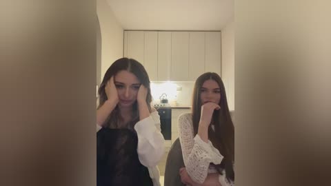 Media: A video shows two young women with long hair, one in a white dress, the other in a knitted sweater, sitting on a couch in a dimly lit living room with beige walls and a white cabinet.