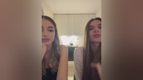 Media: Video of two young women with fair skin and long brown hair, one in a black top, the other in a white lace top, standing in a modern kitchen with white cabinets and a stove.