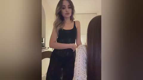 Media: Video of a slender young woman with long blonde hair, wearing a black lace dress, standing in a dimly lit bedroom with a white wall and closed door.