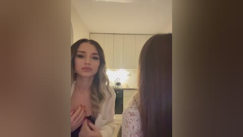 Media: Video of a young woman with fair skin, long wavy hair, and small breasts, wearing a white cardigan, looking at herself in a mirror in a modern, beige-toned bathroom.