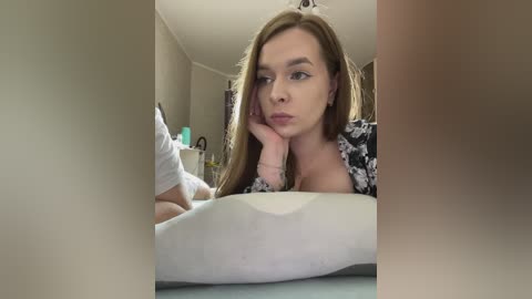 Media: Video of a Caucasian woman with long brown hair, wearing a floral shirt, leaning on a white pillow, looking introspective. Background shows a beige room with a ceiling fan.