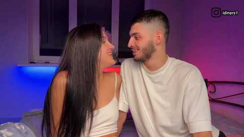 Video of a young couple, a brunette woman in a white tank top and a bearded man in a white t-shirt, sitting close on a bed with purple and blue lighting.