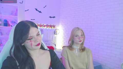 Video of two women in Halloween-themed costumes: one with a black dress and veil, the other in a beige top. Background features white brick wall, bat decorations, and a glowing lamp.
