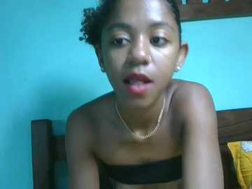 Video of a young Black woman with short curly hair, wearing a black strapless top, gold necklace, and earrings. She's indoors against a teal wall, leaning on a wooden headboard.