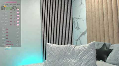 Video of a modern bedroom with a quilted grey bedspread, beige curtains, and a wall featuring vertical black stripes and marble texture.