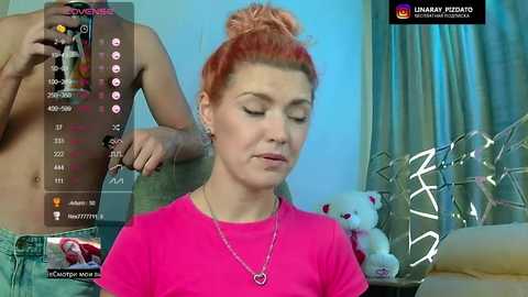 Video of a pale-skinned woman with dyed red hair, wearing a pink shirt, being touched by a topless man in a dimly lit room with teddy bears.