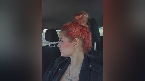 Video of a pale-skinned woman with bright red hair in a messy bun, wearing a black leather jacket and necklace, sitting in a car with dark interiors and blurred scenery outside.