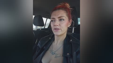 Video of a fair-skinned woman with red hair in a messy bun, wearing a black leather jacket and a heart necklace, sitting in a car with black interior, looking serious.