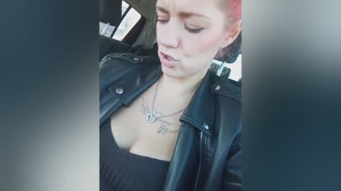 Video of a woman with light skin, short brown hair, wearing a black leather jacket and low-cut black top, revealing cleavage. She has a tattoo on her chest. The background shows the inside of a car with blurry windows.