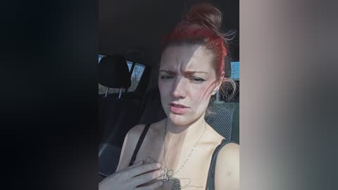 Video of a distressed woman with red hair in a bun, wearing a black top, sitting in a car with dim lighting, looking down.