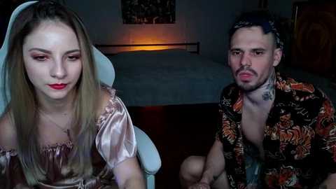 Video of a young woman with long blonde hair, pink satin dress, and a man with a black floral shirt, sitting on a white chair in a dimly lit room.