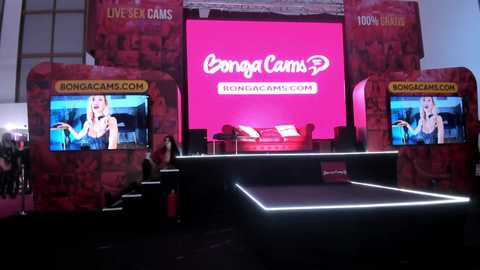 Media: Video of a vibrant, pink-themed event booth featuring \"Bongacam\" branding, with two large screens displaying live chat content. The booth includes a small stage with red couches and a podium.