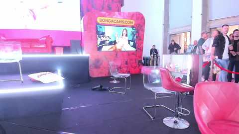 Media: Video of a modern, brightly lit exhibition space with red and pink accents. A large screen displays a woman in a red dress. Clear chairs and red stools are scattered around, with a red couch in the background. People in casual attire mingle.