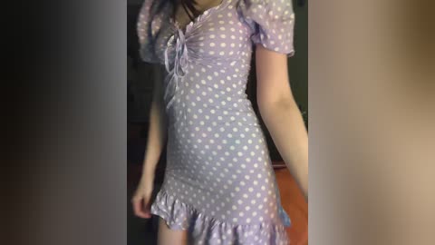 Video of a slender, light-skinned woman in a short-sleeved, light purple polka-dot dress with a ruffled hem, standing indoors with dim lighting.