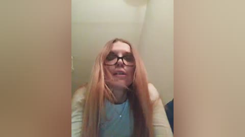 A video of a young woman with long, straight, light brown hair, wearing black-framed glasses and a light blue shirt, standing in a narrow hallway with beige walls.