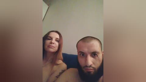 Video of a topless, fair-skinned woman with long red hair and a man with a bald head and beard, both looking into the camera, in a dimly lit room.