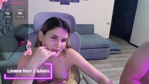 A video of a young woman with light skin and shoulder-length blonde hair, topless, sitting on a grey couch in a modern living room. She has a seductive expression, holding a cherry in her hand.