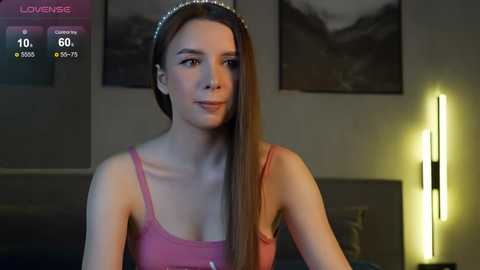 Media: Video of a young woman with straight, light brown hair, wearing a pink tank top, seated in a dimly lit room with a modern lamp and framed art on the wall.