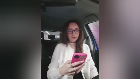 A video of a young woman with straight, dark brown hair and glasses, wearing a white cardigan, taking a selfie inside a car with a pink phone.
