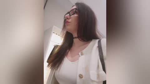 Video of a young woman with long brown hair, glasses, and a white top, standing in a dimly lit, narrow hallway. She has a slender physique and is looking upward.