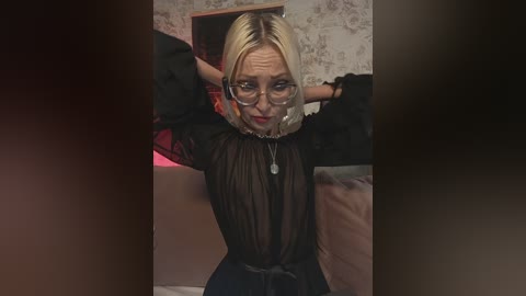 Media: Video of a blonde woman with glasses, wearing a sheer black top and jeans, tied up, in a dimly lit room with floral wallpaper.