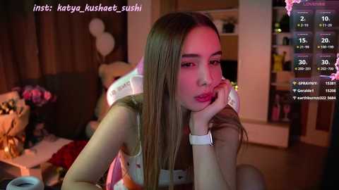 Media: A video of a young woman with long straight brown hair, wearing a white sleeveless top and pink lipstick, leaning on her elbow in a dimly-lit room.