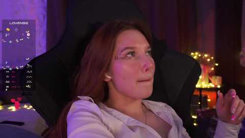 Video of a fair-skinned woman with long auburn hair, wearing a light-colored blouse, sitting in a black gaming chair, illuminated by purple and yellow lights, with a gaming monitor displaying \"LOVING\" in the background.