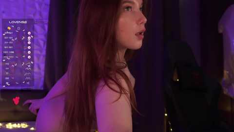 Video of a young woman with long, auburn hair, wearing a beige sweater, in a dimly lit room with purple lighting. She's in profile, looking to the side. Background shows a screen displaying a video game interface.