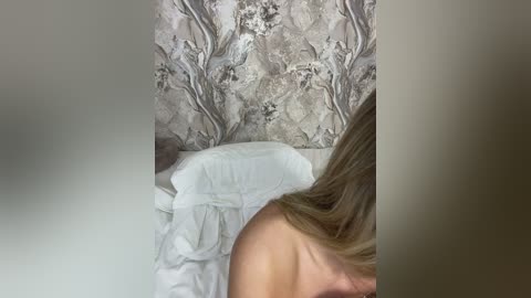 Media: A video captures a partially visible nude woman with light skin and blonde hair, lying on white sheets against a patterned wallpaper background.