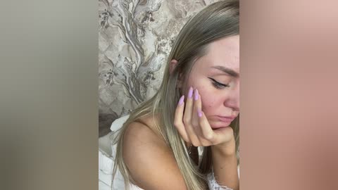 Media: A close-up video of a blonde woman with long hair, wearing pastel-colored nail polish, holding her face with a distressed expression. Background features floral wallpaper and a blurred, beige wall.