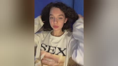 A video of a young woman with curly brown hair, fair skin, and blue eyes, wearing a white \"SEX\" graphic tee, holding her hair back. Background is blurred.